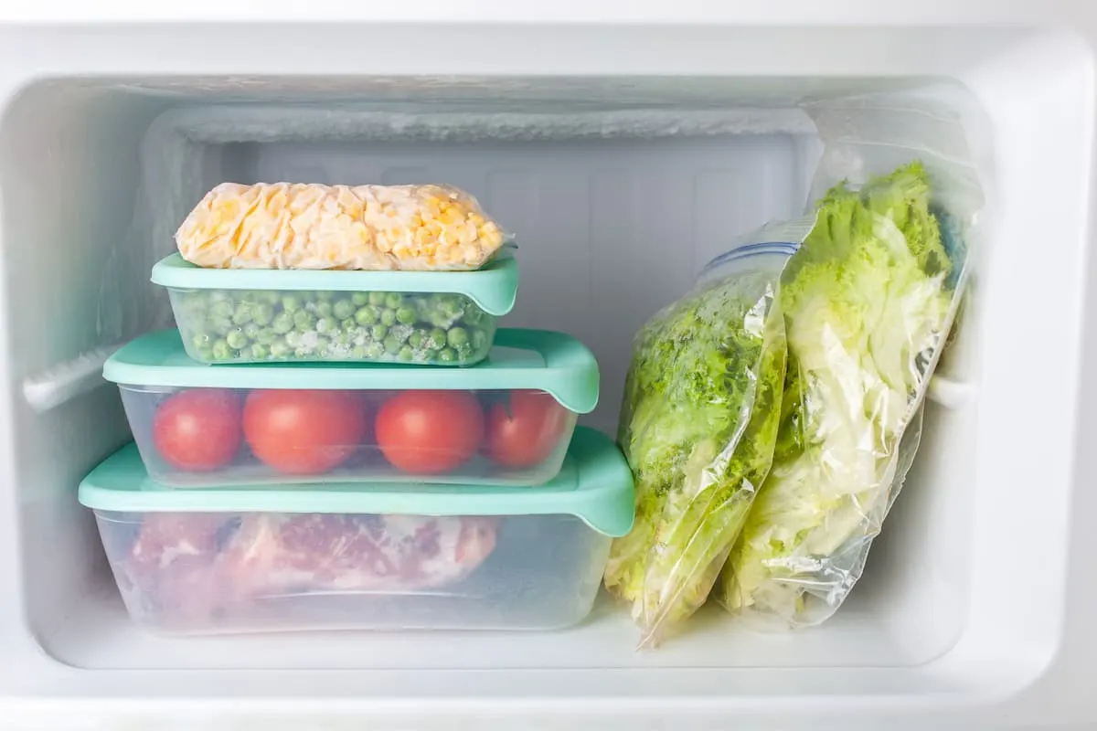 Recipientes de plástico con verduras y alimentos frescos organizados dentro de un refrigerador blanco.
