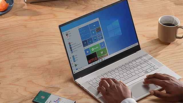 Persona tecleando en una laptop sobre una mesa de madera. Una taza, fruta y un celular están cerca. La pantalla muestra un menú.