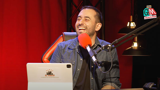 Un hombre ríe frente a un micrófono rojo. Lleva una camisa azul y está sentado en una silla naranja y negra. Hay una laptop y luces.