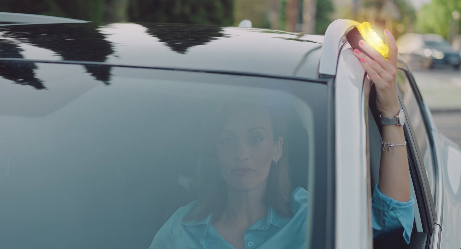 Mujer en coche blanco sostiene luz amarilla. Lleva camisa azul y reloj plateado. Reflejos de árboles en el techo del coche.