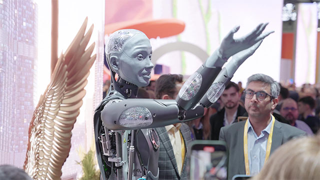 Un robot gris con brazos levantados está en una multitud cerca de una escultura dorada con forma de ala.