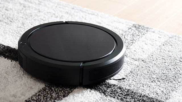 Aspiradora robótica redonda negra sobre una alfombra con diseño geométrico en tonos blancos y grises, contra un fondo de piso de madera clara.