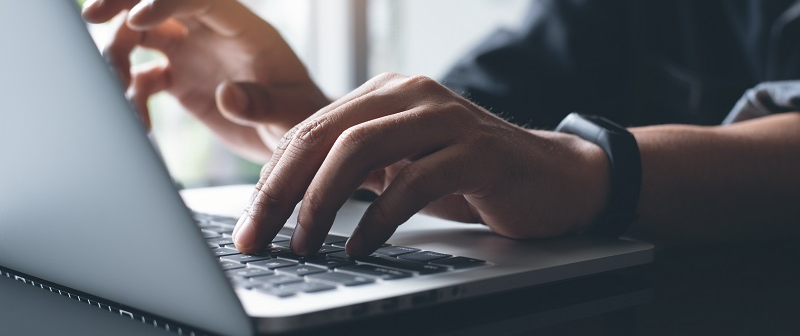 Una persona escribe en un portátil. Se ven las manos sobre el teclado y un reloj en la muñeca. El fondo está borroso.