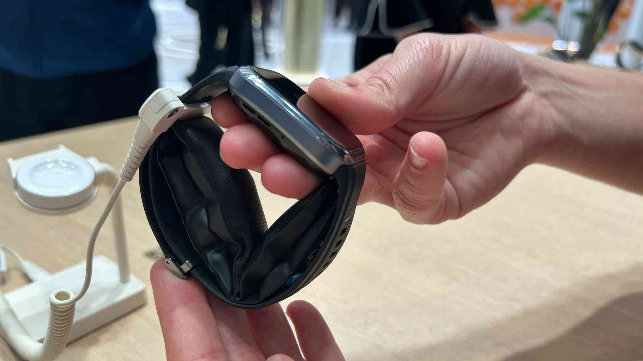 Una mano sostiene un reloj inteligente negro con correa flexible en una mesa clara, cerca de un soporte de exhibición blanco.