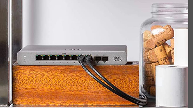 Un router gris de Cisco está sobre un bloque de madera. Cables negros están conectados al router. Un bote de cristal con corchos está cerca.