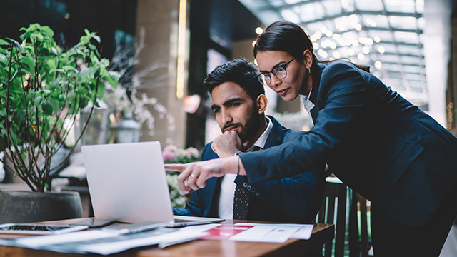 Un hombre y una mujer con trajes azules ven un portátil sobre una mesa de madera con documentos. Hay una planta cerca.