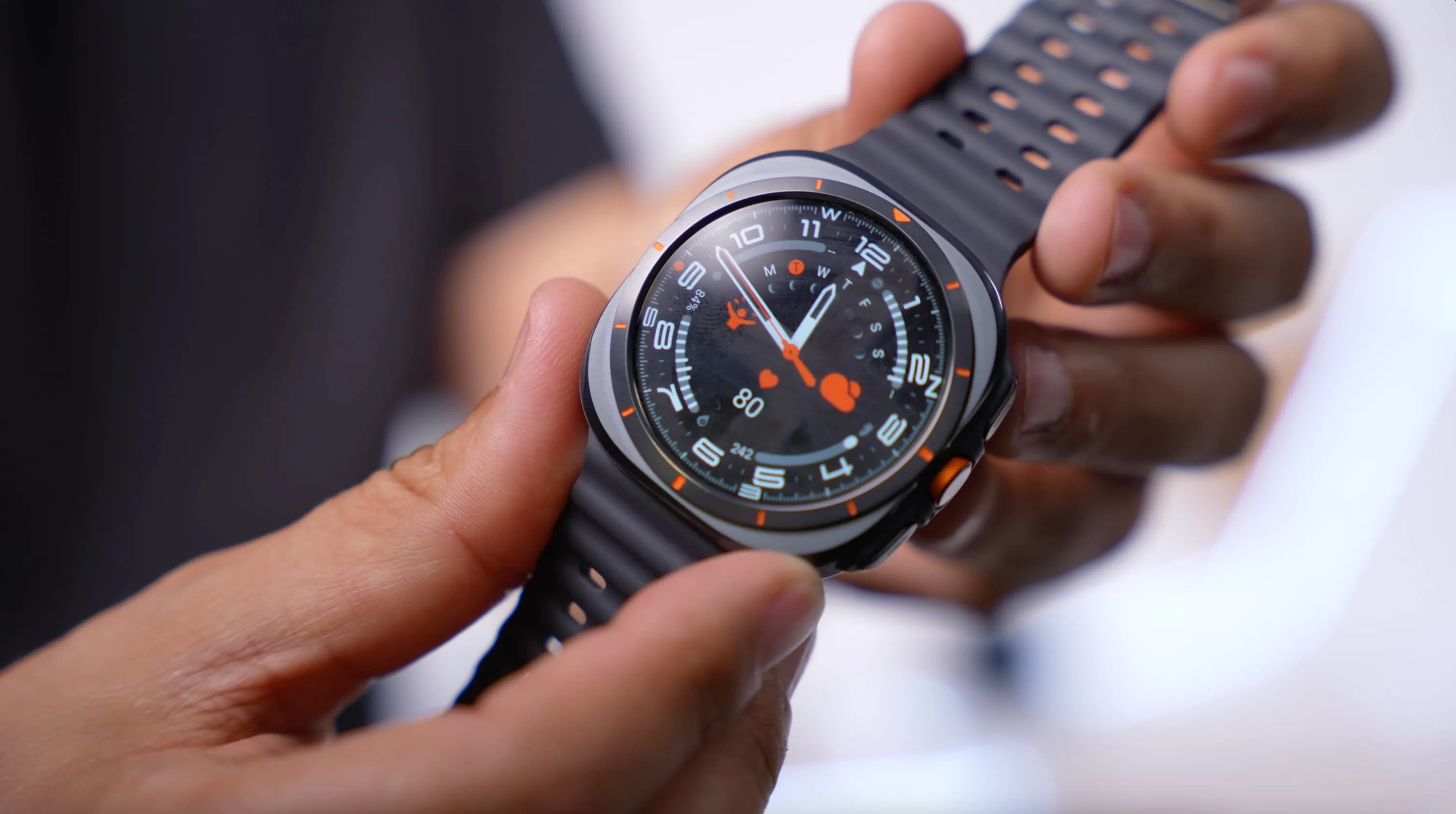 Un reloj inteligente con esfera negra y detalles naranjas es sostenido por una mano; la correa es gris oscura con perforaciones.