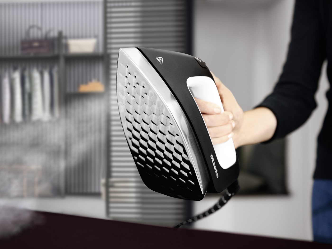 Mano sujetando una plancha vertical negra y plateada. Al fondo, un armario con ropa y estantes.