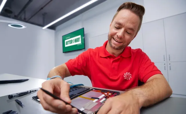 Un hombre con camisa roja repara un dispositivo electrónico sobre una mesa gris, con herramientas y un letrero verde en la pared.