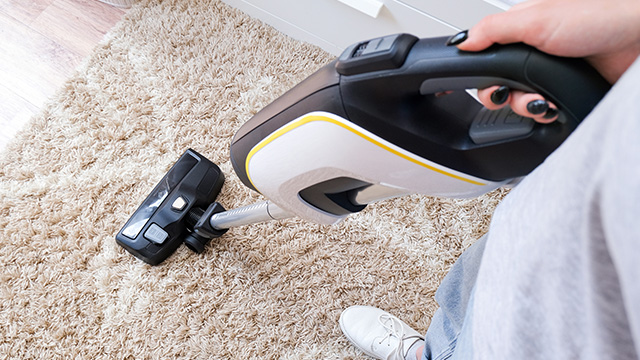 Una persona aspira una alfombra con una aspiradora vertical blanca, negra y amarilla. Se ve un pie con un zapato blanco.