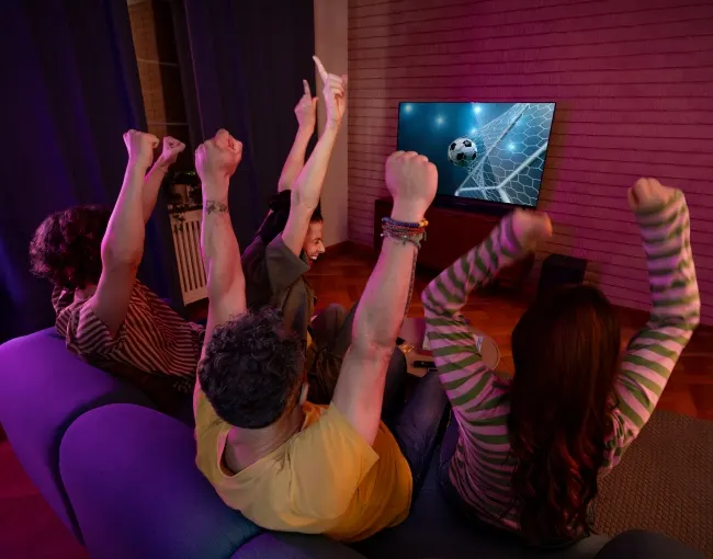 Un grupo de amigos vitorean un partido de fútbol en la televisión con los brazos en alto, sentados en un sofá morado.