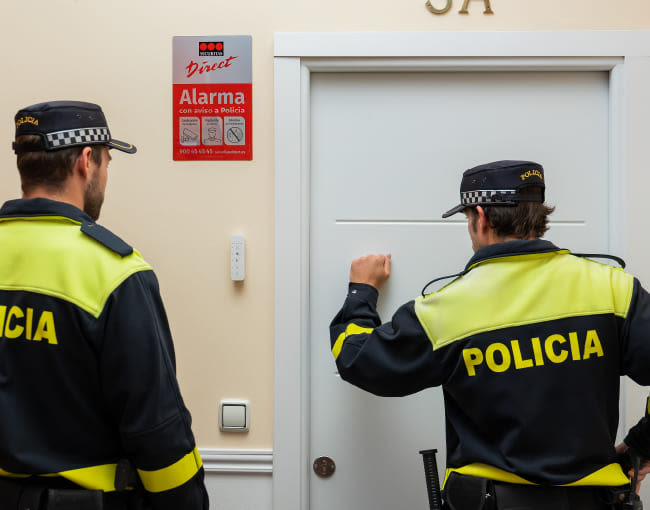 Dos vigilantes de seguridad con uniforme azul y chaleco naranja, vistos de espaldas, llaman a una puerta blanca interior bajo cartel rojo de alarma conectada.