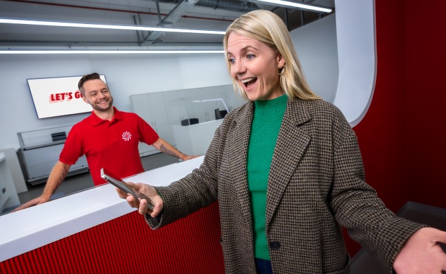 Mujer sonriente con un teléfono en un mostrador rojo y blanco. Empleado con camisa roja al fondo.
