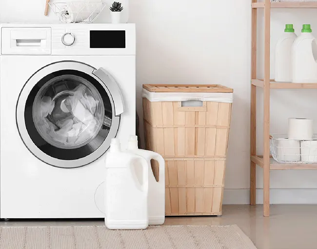 Lavadora blanca con ropa dentro, junto a un cesto de ropa de madera y un estante con productos de limpieza y papel higiénico.