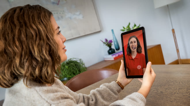 Persona realiza videollamada en una tablet con una asesora de tecnología desde su casa, sentada a la mesa del comedor, recibiendo asesoramiento.