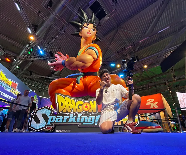 Un hombre arrodillado sostiene una cámara frente a un cartel de "Dragon Ball" con una figura de Gokú y el logo de "2K".