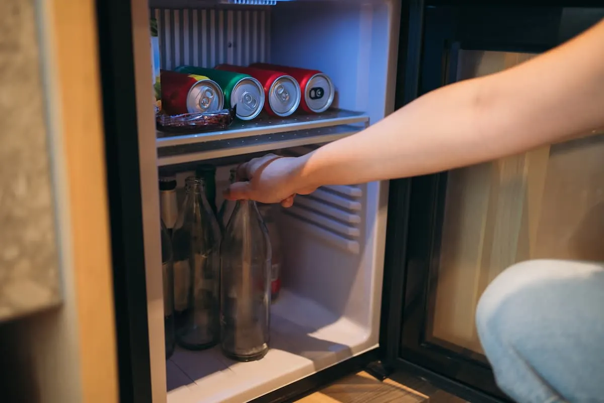 Persona sacando una botella de vidrio de una nevera pequeña con latas en el estante superior.