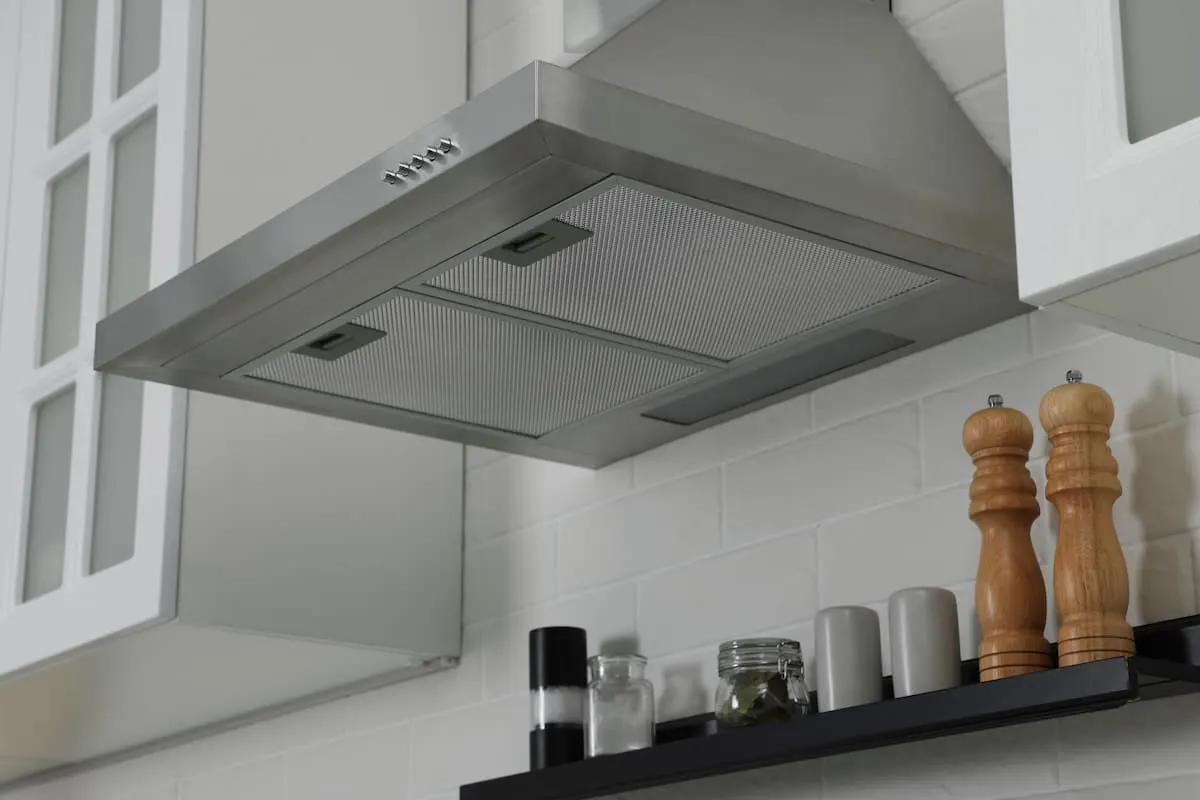 Extractor de cocina de acero inoxidable con armarios blancos y una repisa con especias y molinillos de madera.