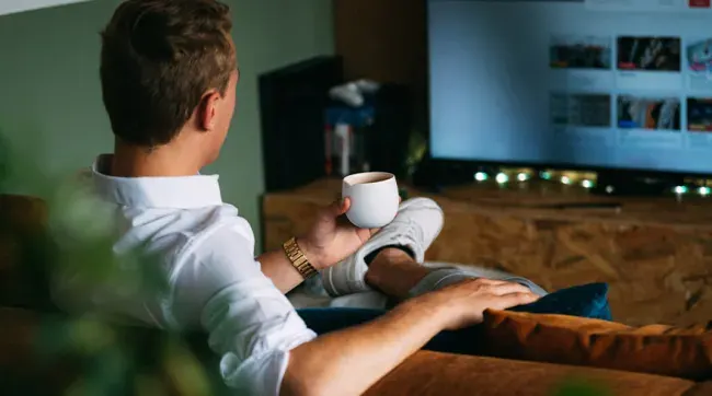 Un hombre en camisa blanca está sentado en un sofá marrón, viendo la televisión y sosteniendo una taza blanca.