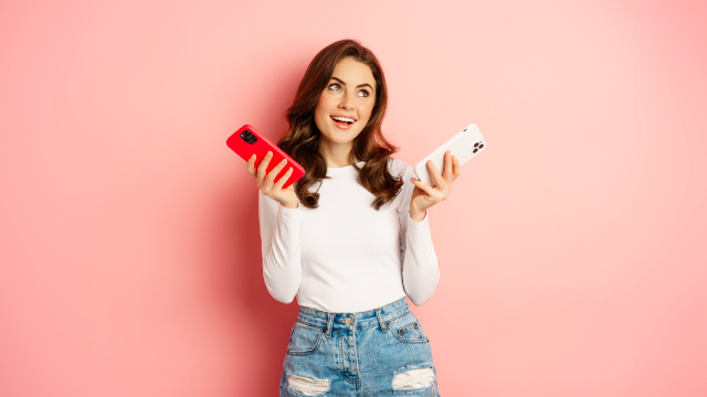 Mujer con camisa blanca y jeans azules sostiene un teléfono rojo y otro blanco sobre un fondo rosa.