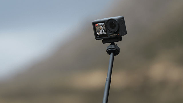 Cámara negra en un palo negro con una pantalla activa sobre un fondo borroso azul y marrón.