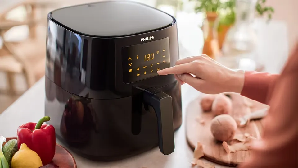 Freidora negra Philips con panel digital marcando 180 grados, mano ajustando la configuración, verduras y tabla de cortar con alimentos.