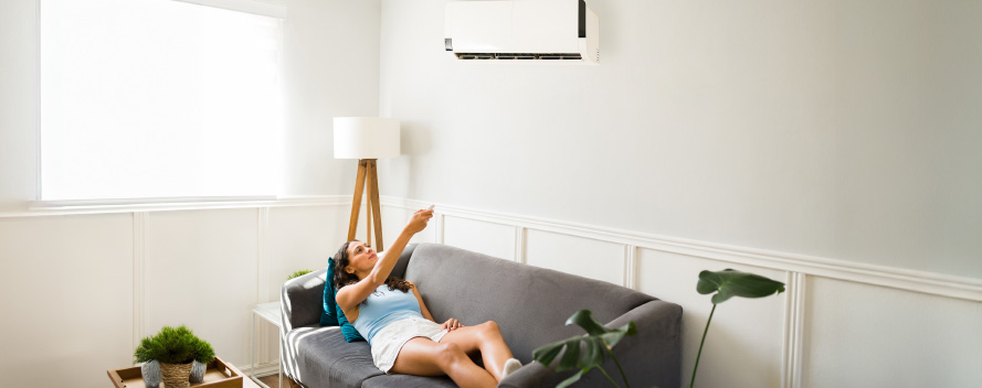 Mujer en sofá gris apunta control remoto al aire acondicionado blanco en pared blanca. Lámpara, ventana y planta visibles.