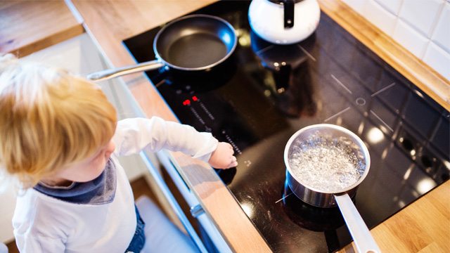 Vista elevada de un niño cerca de una estufa con una sartén, tetera blanca y una olla con agua hirviendo.