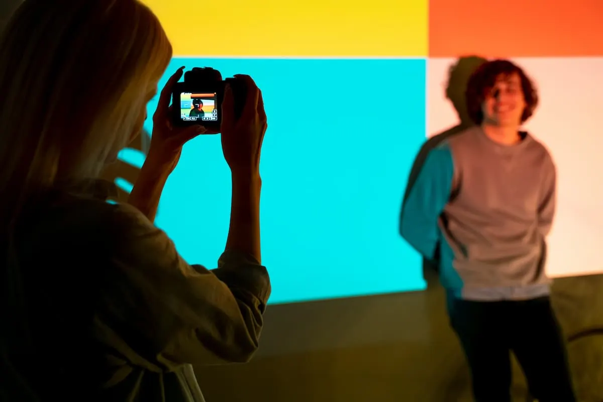 Persona tomando una foto con cámara a un joven posando frente a una pared iluminada con colores brillantes.