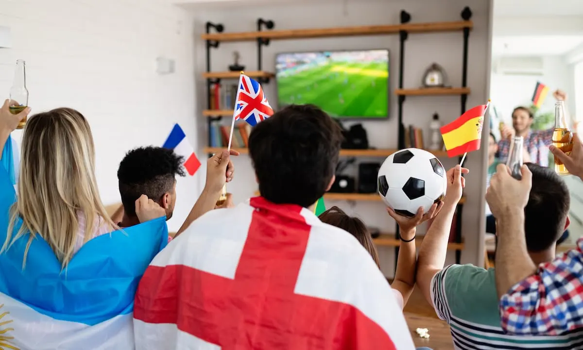 Grupo de amigos celebrando con banderas de varios países mientras ven un partido de fútbol en televisión.