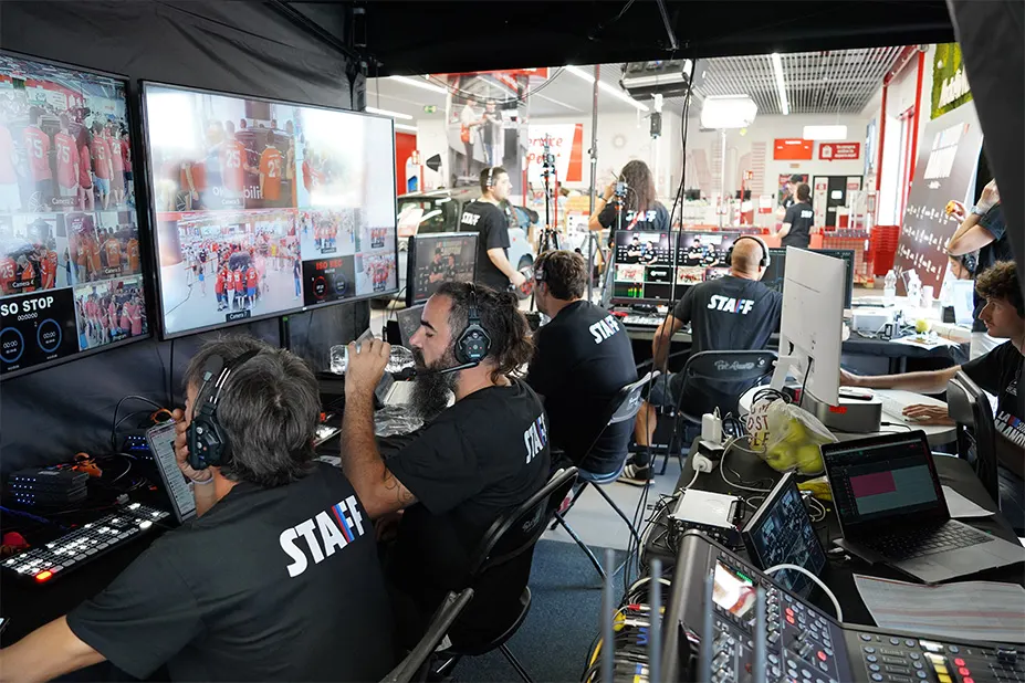 Equipo de producción de televisión en sala con monitores, ordenadores y personal con auriculares y camisetas negras.