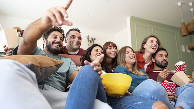 Grupo de personas sonriendo en un sofá con palomitas y comida para llevar. Un hombre señala hacia arriba.