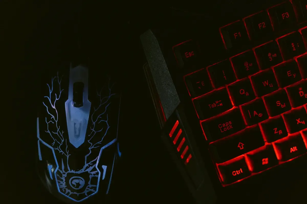 En un fondo oscuro, un ratón con líneas azules y un teclado con retroiluminación roja están uno al lado del otro.