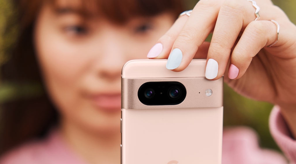 Una mujer sostiene un teléfono móvil color melocotón con lentes de cámara negras y un flash. Sus uñas están pintadas de colores pastel.