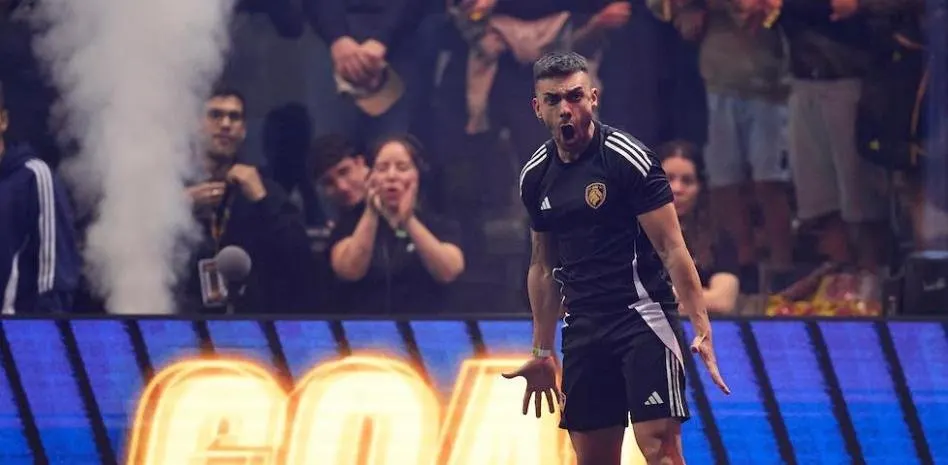 Hombre con uniforme deportivo negro y blanco celebra frente a público y pantalla azul brillante con letras doradas. Humo visible.
