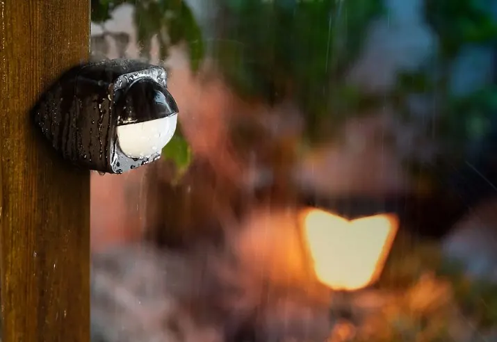 Sensor de movimiento exterior bajo la lluvia, instalado en un poste de madera.