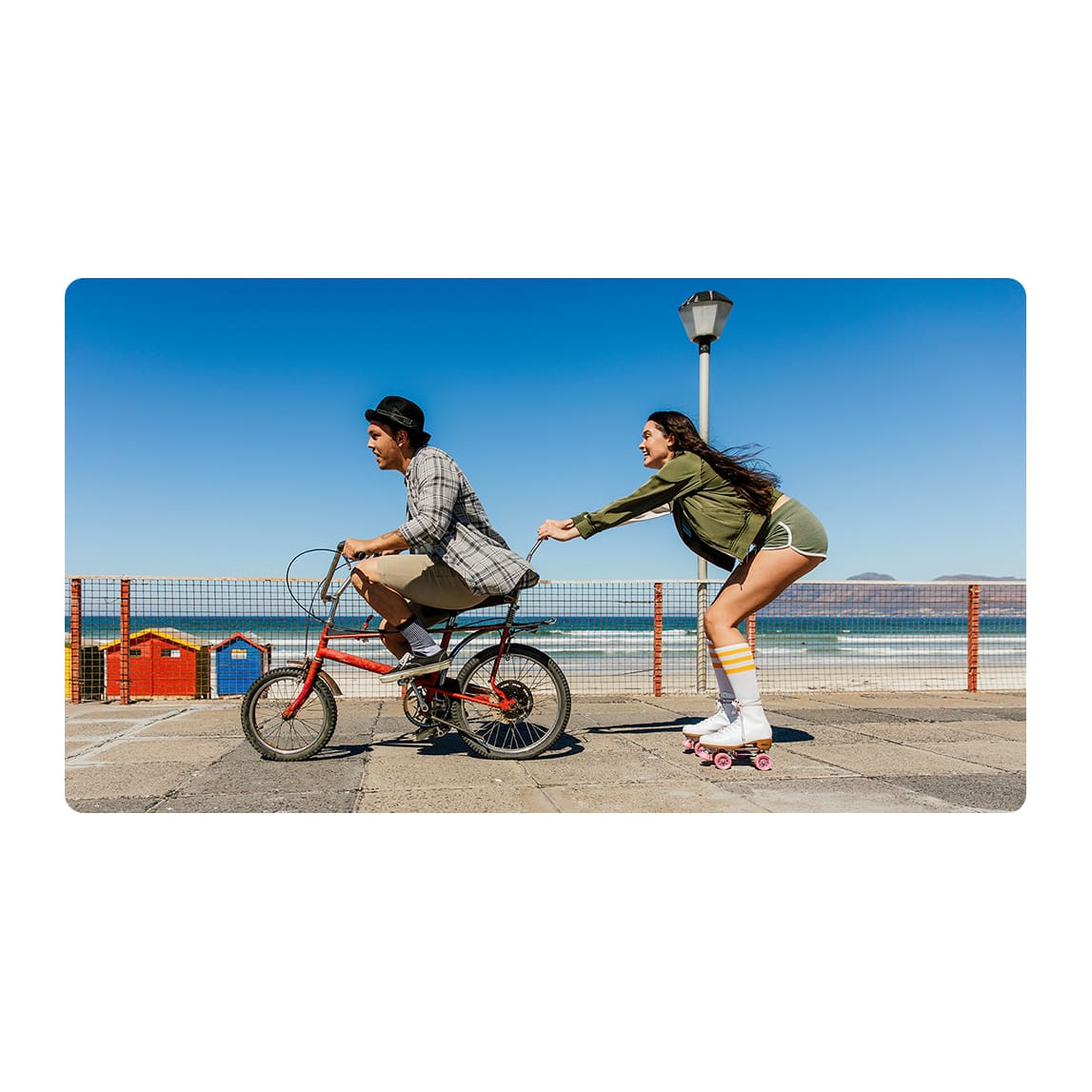 Un hombre en bicicleta roja es empujado por una mujer en patines blancos cerca de la playa, bajo un cielo azul claro.