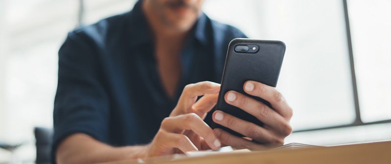 Un hombre usa un móvil negro. Lleva una camisa azul oscura. La ventana blanca está al fondo.