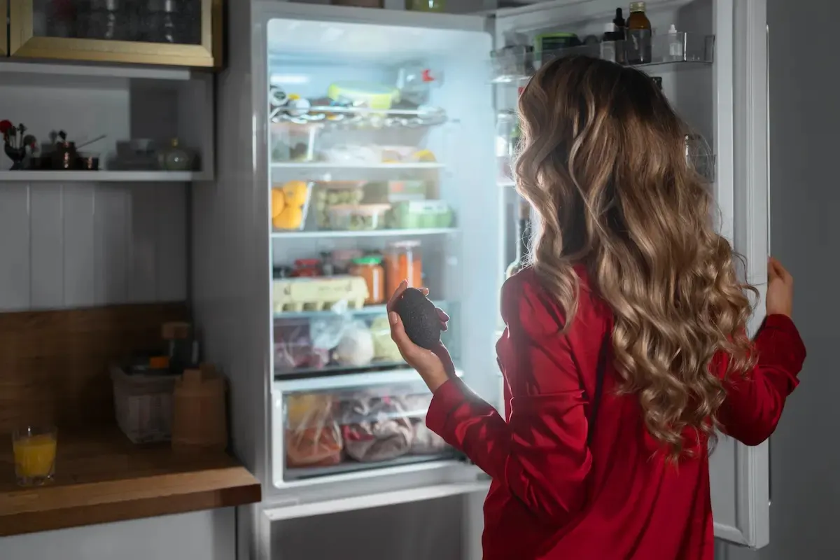 Una persona abre un refrigerador lleno de comida, sosteniendo un alimento en la mano, en una cocina con estantes y encimera.