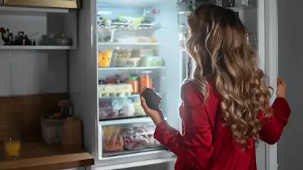 Una persona abre un refrigerador lleno de comida, sosteniendo un alimento en la mano, en una cocina con estantes y encimera.