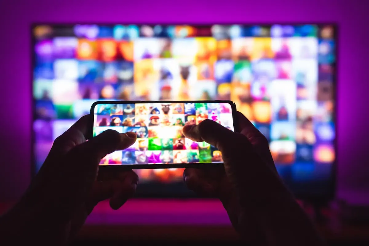 Manos sosteniendo un smartphone frente a un televisor con pantalla de streaming