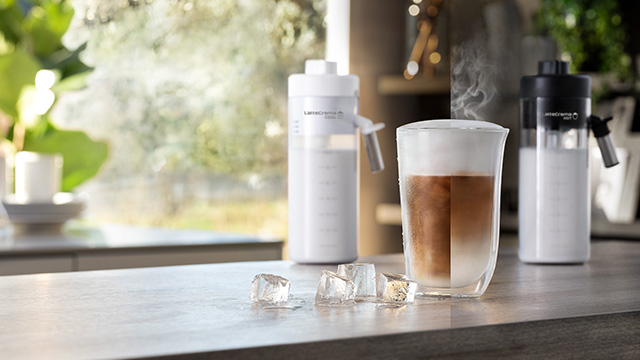 Vaso con café espumoso, hielos, y dos botellas blancas sobre una mesa gris. Al fondo, se ve una ventana con vegetación.