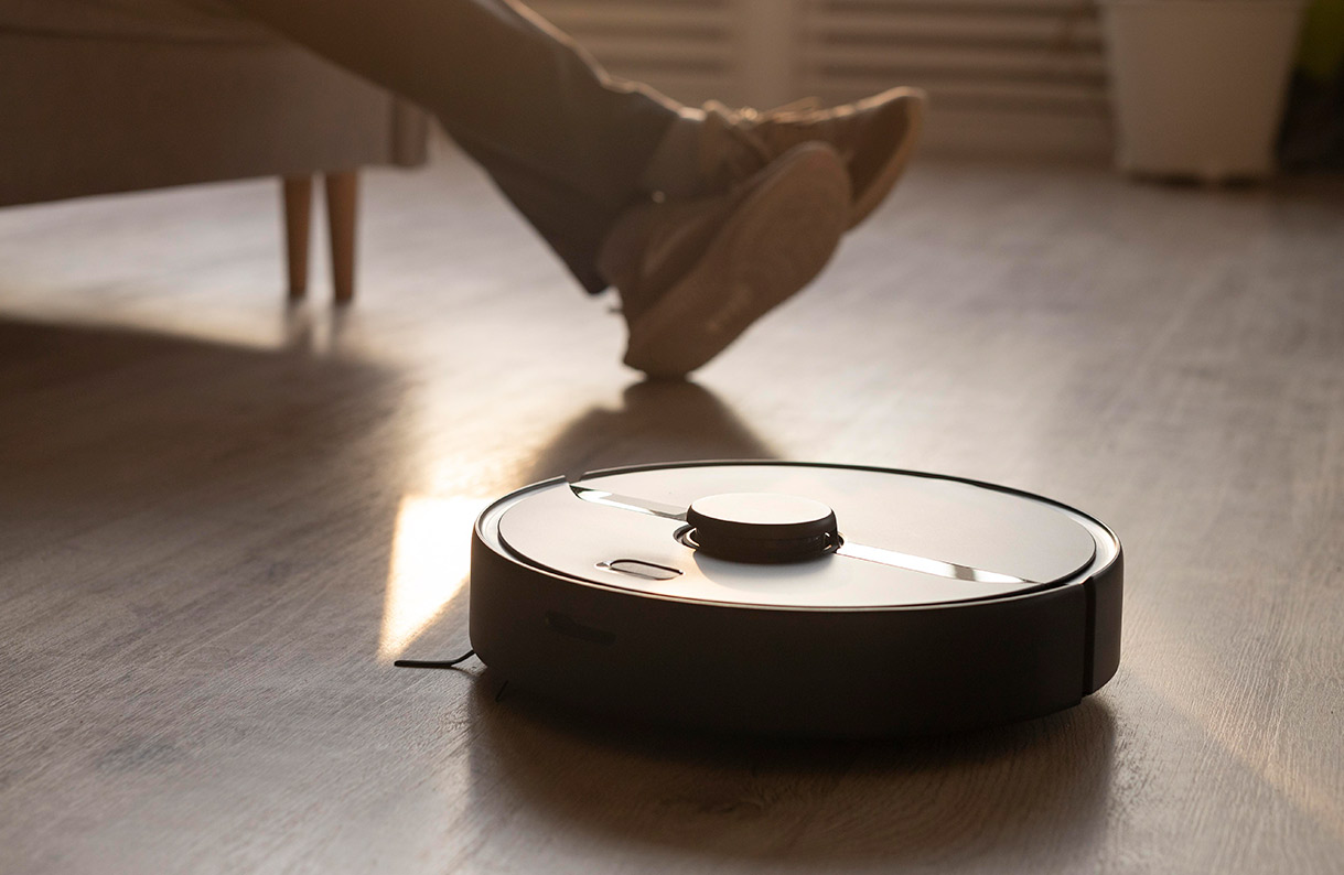 Un robot aspirador redondo, negro y blanco, está en un piso de madera, con pies en zapatos marrones en segundo plano.