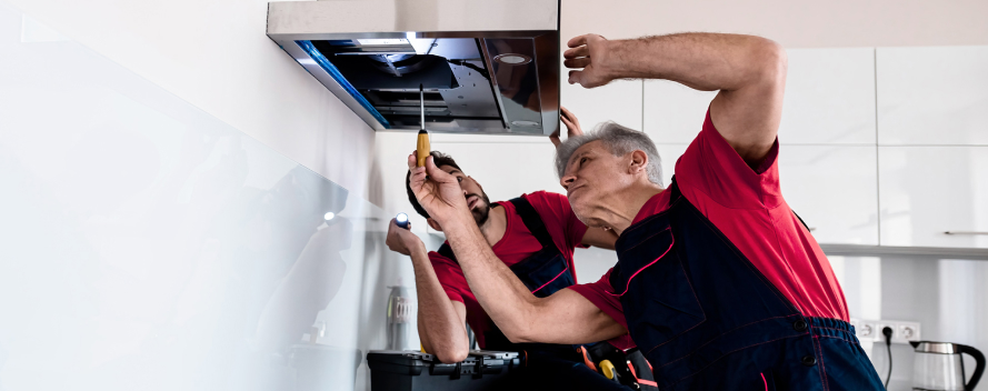 Dos hombres reparan una campana extractora en una cocina. Uno usa un destornillador, el otro sostiene una linterna.