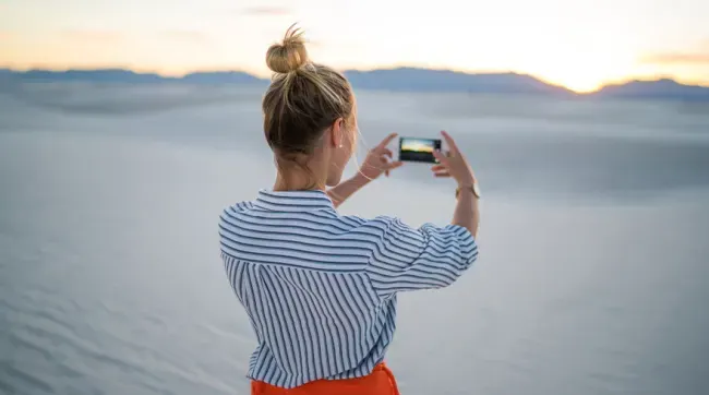 Mujer de espaldas tomando foto con móvil. Lleva camisa a rayas azules y blancas, falda naranja y moño. Fondo de arena y cielo al atardecer.
