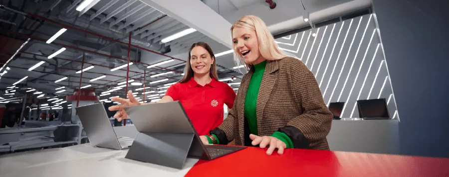 Dos mujeres de pie frente a portátiles en una mesa roja. La de la izquierda lleva una camisa roja.