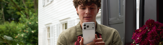 Joven mostrando iPhone blanco en sus manos ante la cámara, en exterior con puerta gris y flores rojas de fondo.