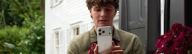 Joven mostrando iPhone blanco en sus manos ante la cámara, en exterior con puerta gris y flores rojas de fondo.