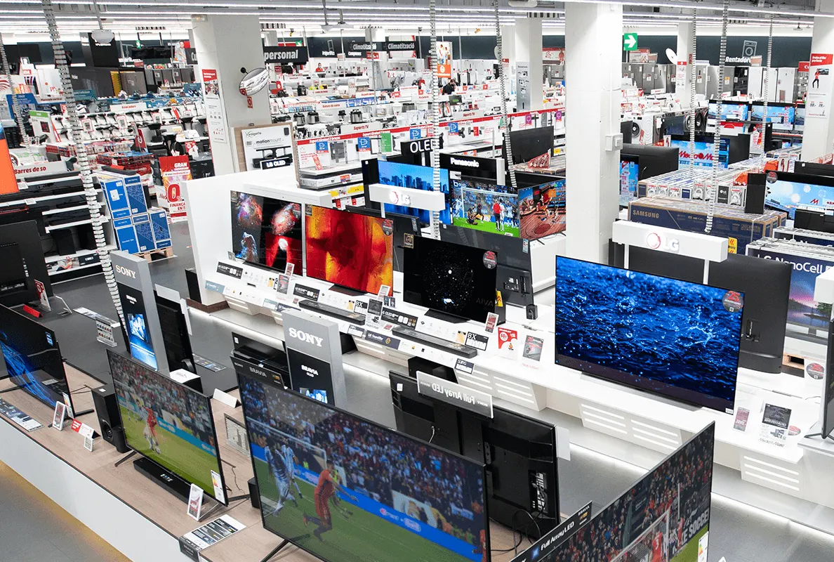 Interior de tienda con televisores en exhibición sobre estantes blancos. Se ven otros productos al fondo y letreros colgantes.