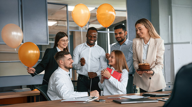 Un grupo de personas celebra un cumpleaños en una oficina con globos naranjas, un pastel y un regalo rojo.
